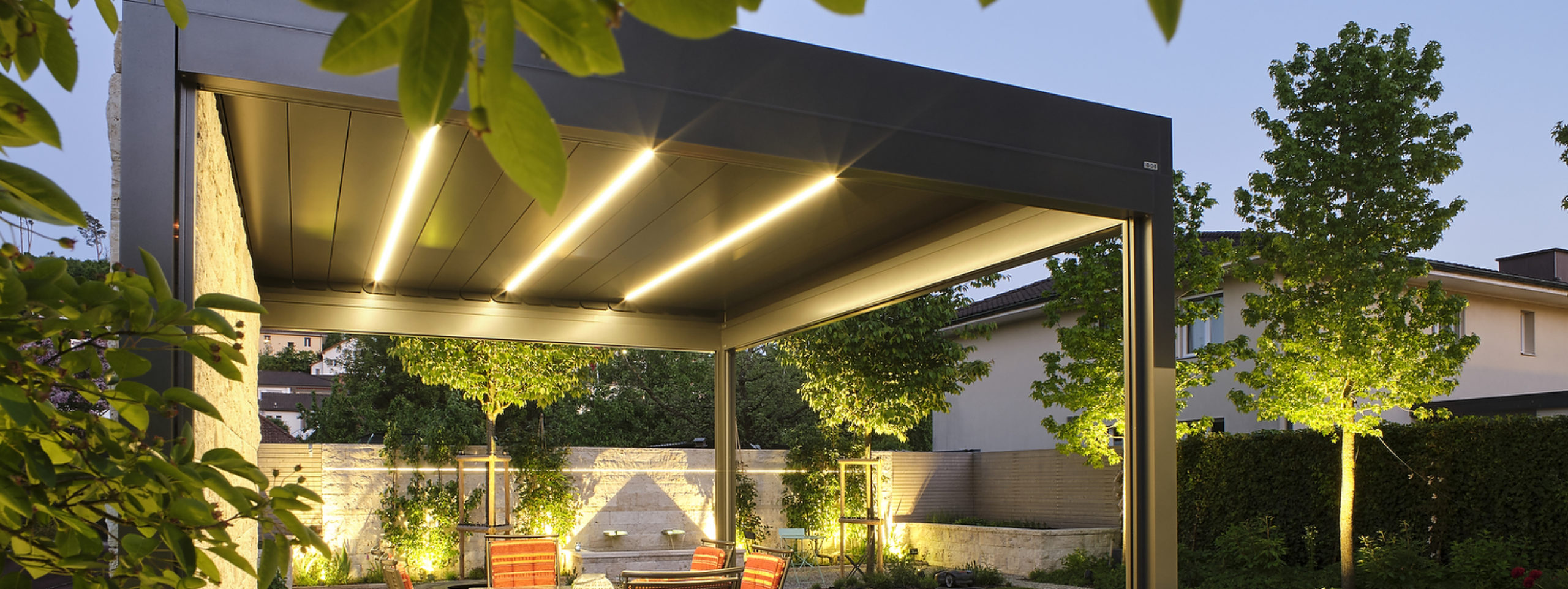 Das Lamellendach ist ein perfekter Schutz gegen Sonne und Regen. Die natürliche Luftzirkulation durch die Lamellen funktioniert wie eine umweltfreundliche Klimaanlage. Bei Sonnenschein stellt man die Lamellen einfach so schräg, damit man den nötigen Schatten und die gewünschte Luftzirkulation erhält. Bei Schlechtwetter schliesst man die Lamellen, um vor Regen geschützt zu sein. Auf dem Bild ist das Lamellendach Bavona von Sunparadies mit integrierten LED-Strips in der sommerlichen Abenddämmerung zu sehen.